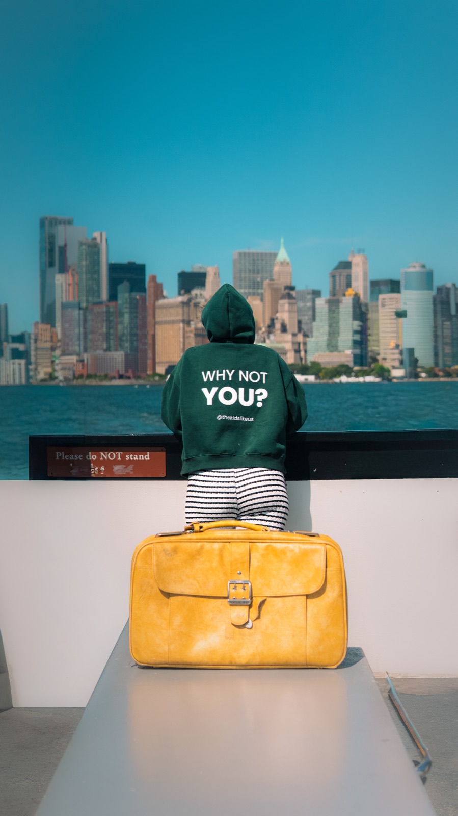 Someone in the Why Not You? hoodie looking out at the New York skyline beside the yellow suitcase