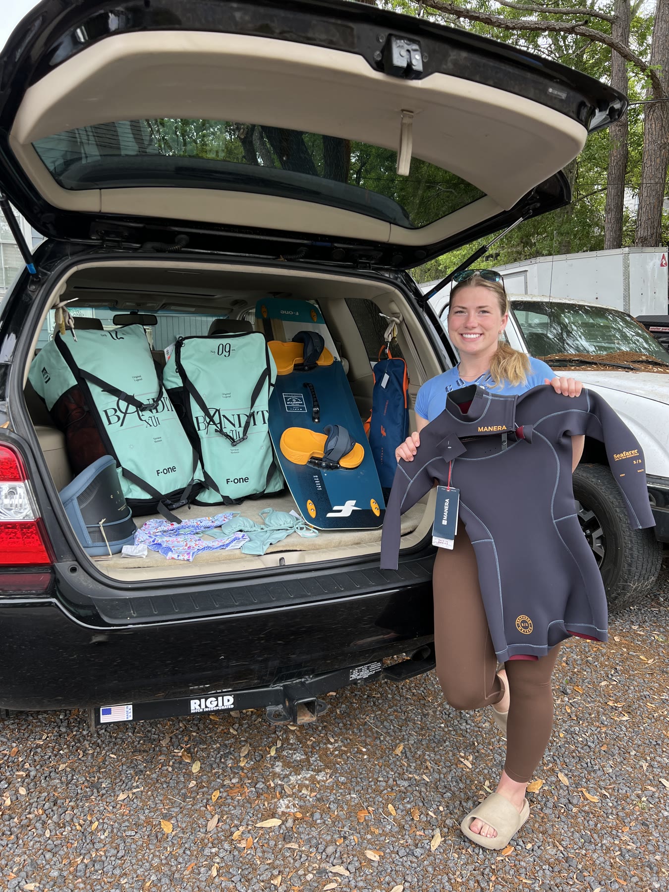 Syd Meyer holding up a new wetsuit beside a car trunk full of sponsor kites and boards